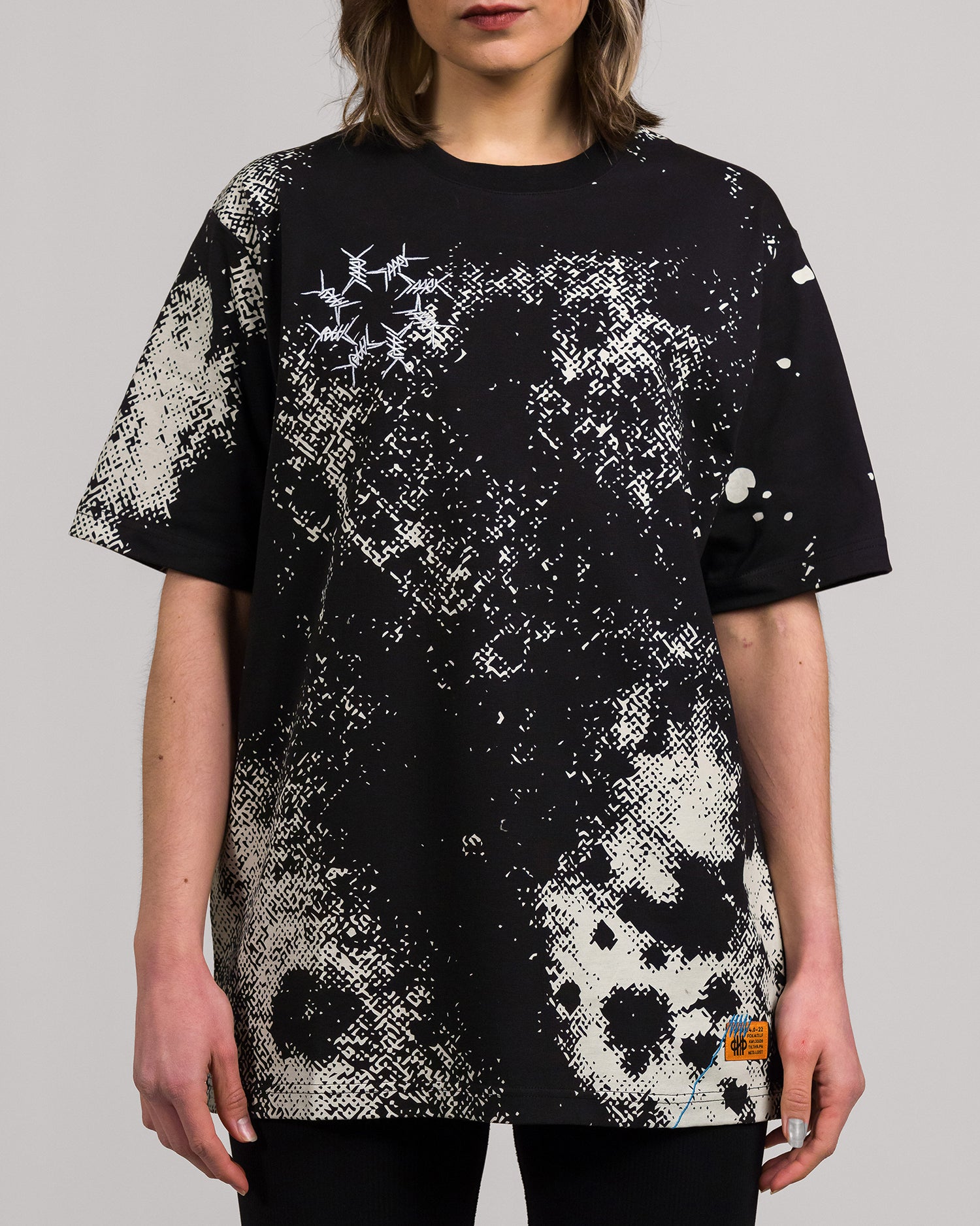 Ein schwarzes Oversize-T-Shirt mit abstraktem, pixelartigem Muster in Beige und Weiß auf der Vorderseite. Die linke Brust zeigt ein weißes, grafisches Motiv aus stilisierten Stacheldrahtlinien. Rechts unten ist ein kleines, rechteckiges Label in Orange mit Logo und Text aufgenäht.