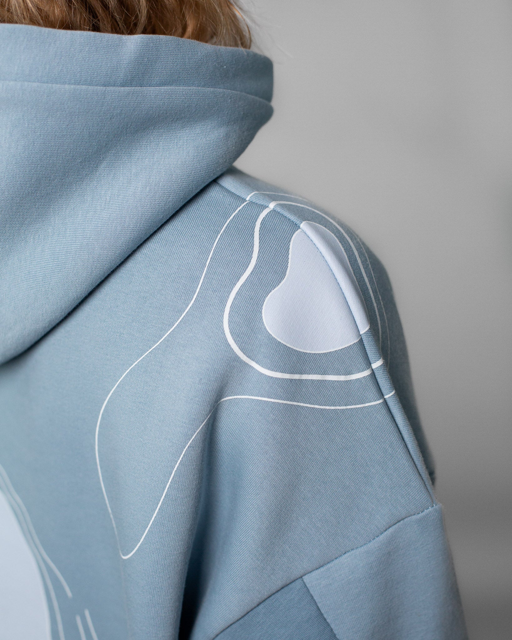 Detailaufnahme eines hellblauen Kapuzenpullovers. Zu sehen ist die rechte Schulterpartie mit einem weißen, organisch verlaufenden Linienmotiv, das sich über den Stoff zieht. Der Fokus liegt auf der glatten Stoffstruktur, der präzisen Nahtführung und dem grafischen Print, der an topografische Formen erinnert.