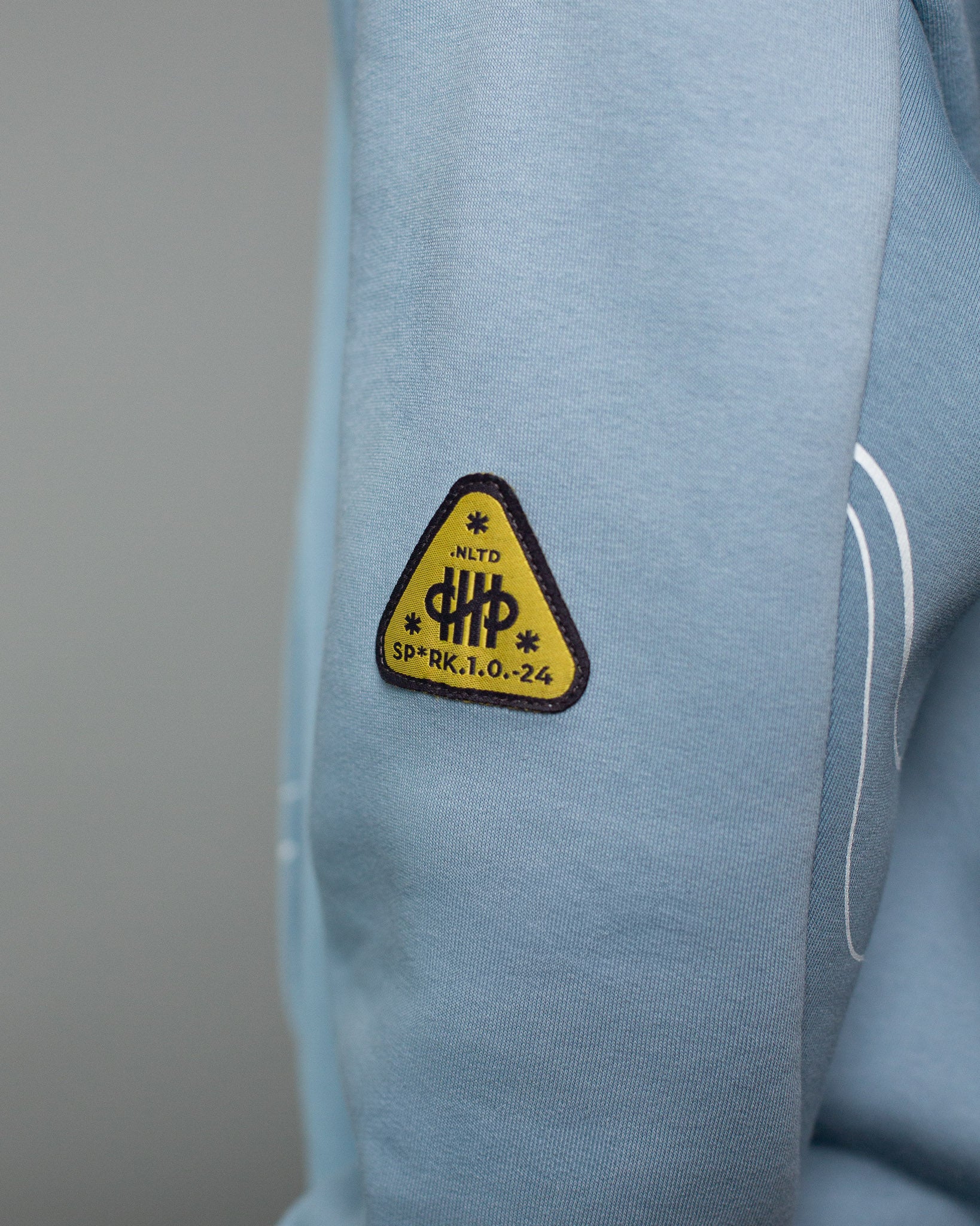 Detailaufnahme eines hellblauen Kapuzenpullovers. Auf dem linken Ärmel befindet sich ein dreieckiger Patch in Gelb mit schwarzer Umrandung und Aufschrift „SP*RK.1.0.24 – NLTD“. Der Aufnäher zeigt ein stilisiertes Symbol und ist von feinen weißen Liniengrafiken umgeben. Der Fokus liegt auf dem markanten Ärmelpatch als Designdetail.