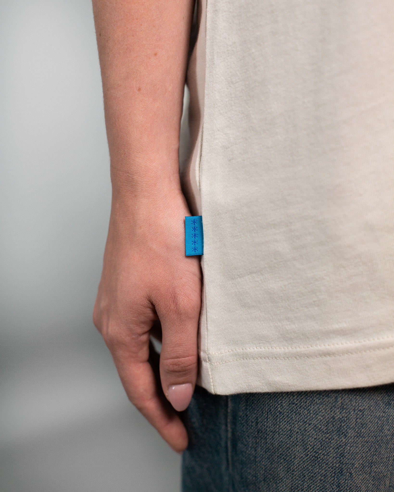 Detailaufnahme des unteren Saums eines beigefarbenen T-Shirts. An der Seitennaht ist ein kleines, leuchtend blaues Stofflabel mit vier Sternsymbolen angenäht. Der Fokus liegt auf dem dezenten Labeldetail und der feinen Stoffstruktur des T-Shirts.