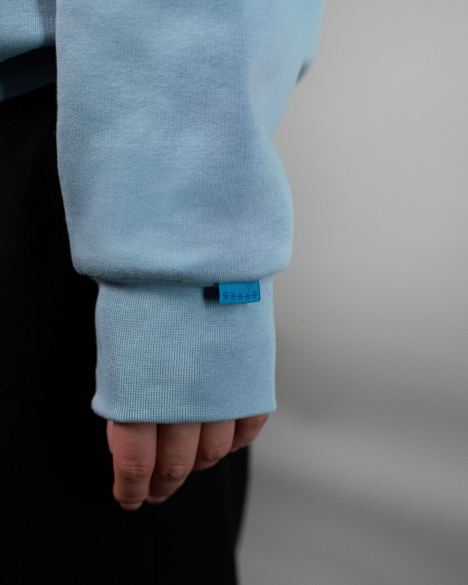 Detailaufnahme des Ärmels eines hellblauen Kapuzenpullovers. Am Bündchen ist ein kleines blaues Stofflabel mit vier Sternsymbolen angenäht. Der Fokus liegt auf dem feinen Labeldetail und der weichen Stoffstruktur des Hoodies. Der Hintergrund ist neutral und unscharf gehalten.