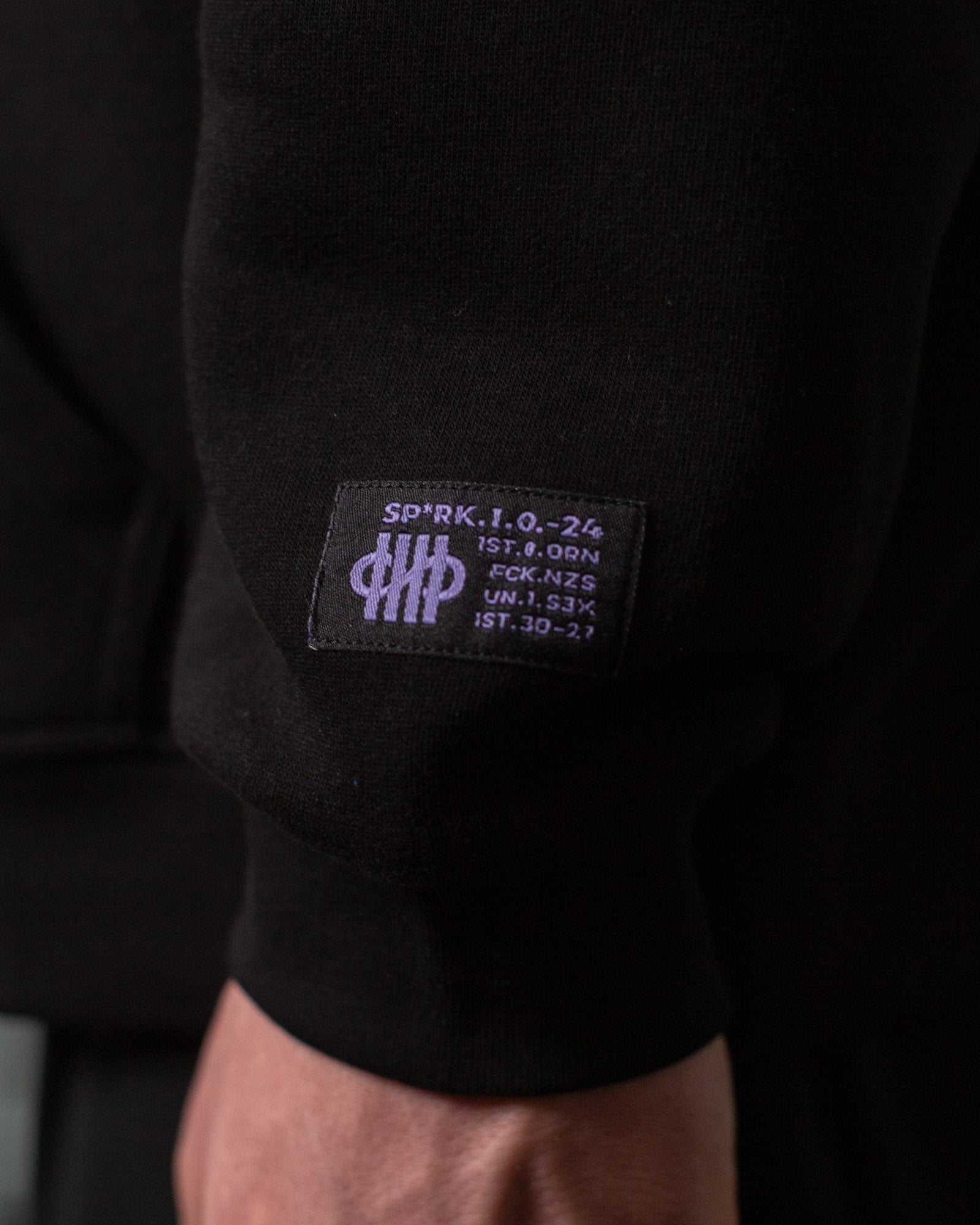Nahaufnahme des Ärmelpatches unten rechts am Hoodie. Auf dem schwarzen Label befindet sich ein lilafarbenes Emblem mit stilisierter Zählgrafik sowie technische Anmutung durch kryptische Zeichen wie „SP*RK.I.O.-24“ und weitere Codezeilen.