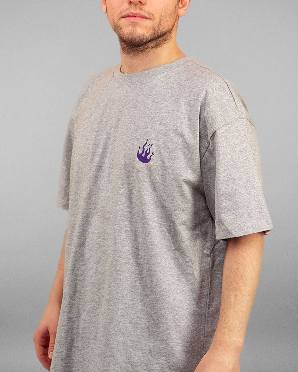 Eine Person trägt ein graues T-Shirt mit kleinem, violettem Flammenmotiv auf der linken Brustseite. Innerhalb der Flamme sind kleine Buchstaben angeordnet. Das Shirt hat eine lockere Passform mit überschnittenen Schultern und Rundhalsausschnitt. Der Fokus liegt auf dem minimalistischen Frontdesign mit der violetten Flamme.