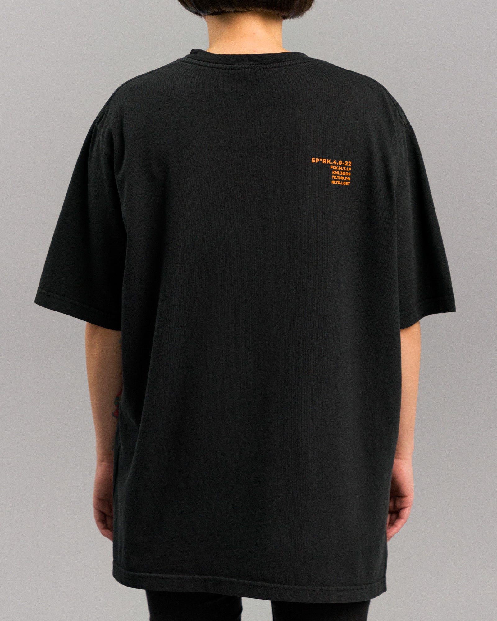 Rückseite eines schwarzen, locker sitzenden T-Shirts mit kleinem orangen Textprint im rechten Schulterbereich. Der Print enthält codierte Begriffe und Zahlen. Die Ärmel sind weit geschnitten.