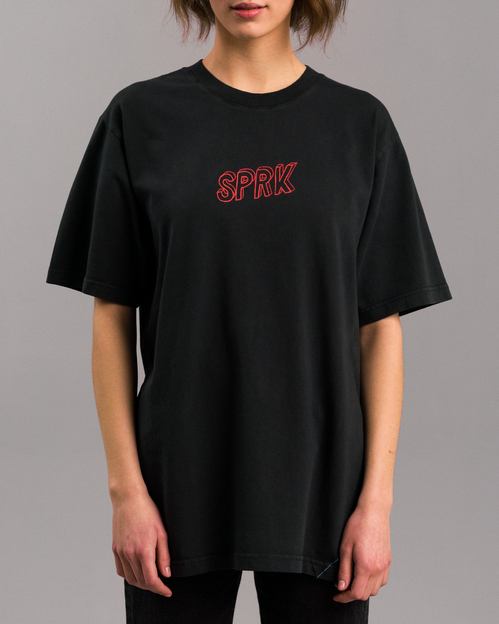 Frontalansicht einer Person, die ein locker geschnittenes schwarzes T-Shirt mit rotem „SPRK“-Schriftzug auf der Brust trägt.
