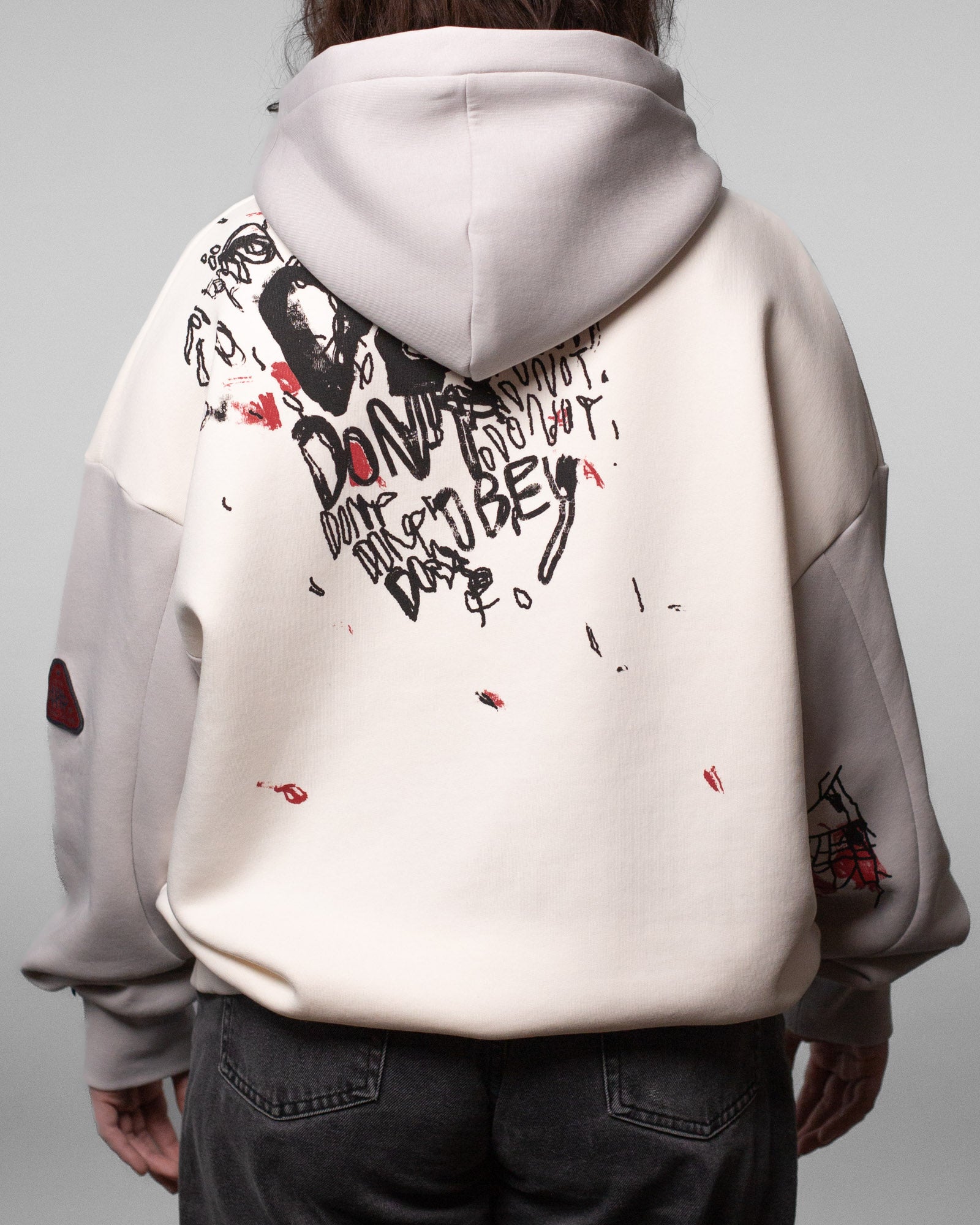 Rückansicht des Hoodies mit auffälligem schwarz-rotem Rückendruck. Der Print zeigt Kritzelgrafiken, Gesichter und den handschriftlich wirkenden Schriftzug „DON’T OBEY“ mehrfach wiederholt. Auf dem Ärmel sind rote und schwarze Linienzeichnungen sichtbar.