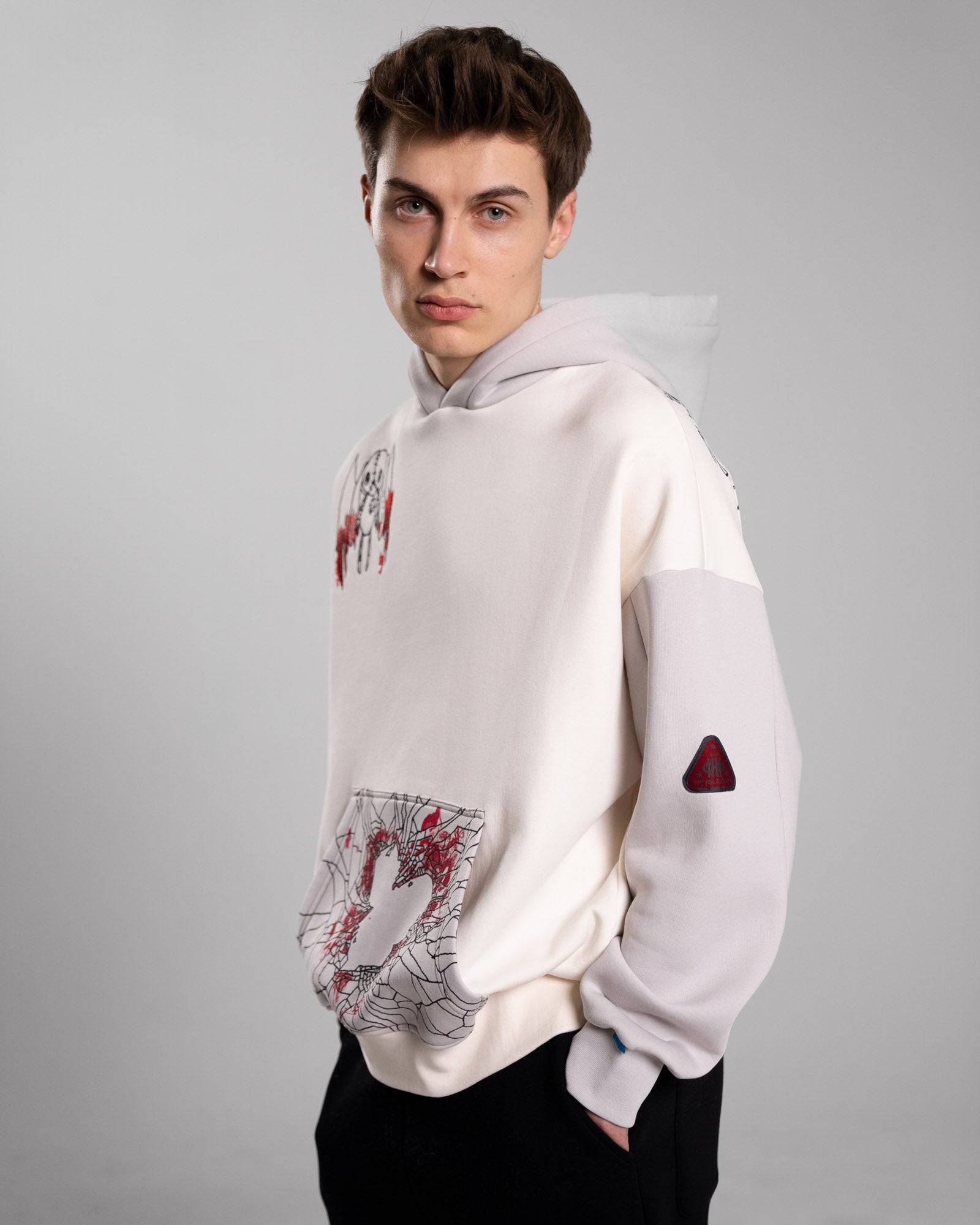 Seitenansicht eines männlichen Models im Hoodie. Gut sichtbar sind das rote Ärmelpatch, die Brustillustration der geflügelten Figur und die grafisch gestaltete Kängurutasche mit Herzumriss.