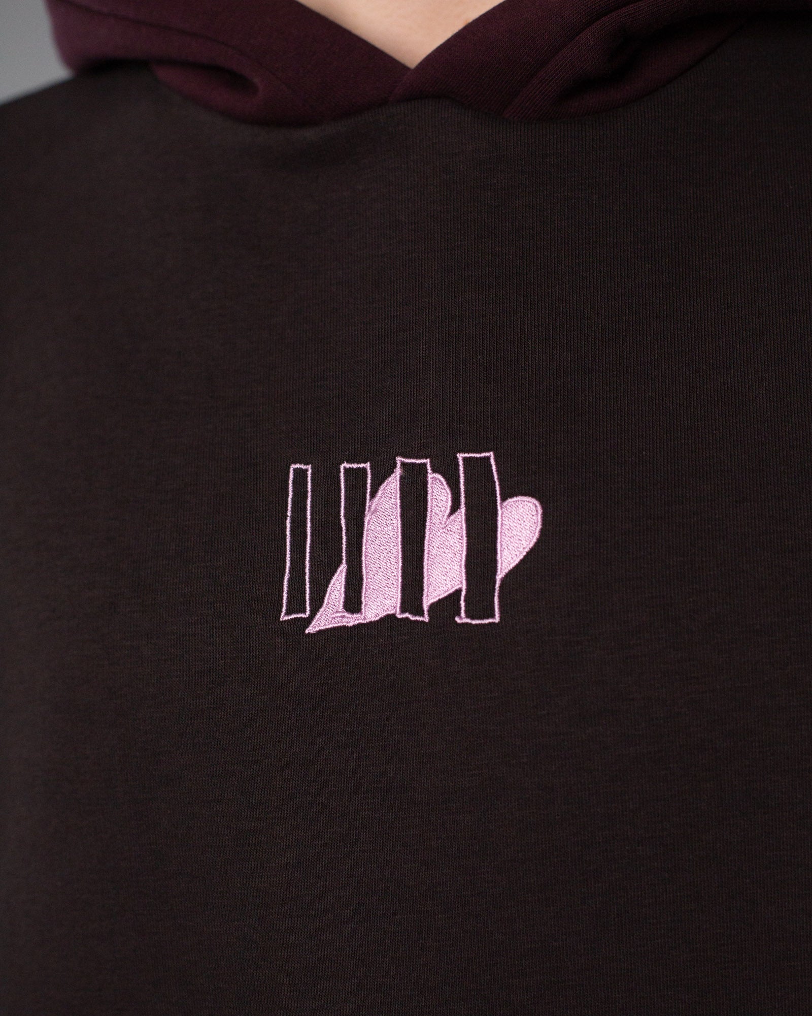 Detailaufnahme des Brustlogos: fünf senkrechte Linien mit integrierter Herzform, aufgestickt in hellem Rosa auf dunkelbraunem Stoff.