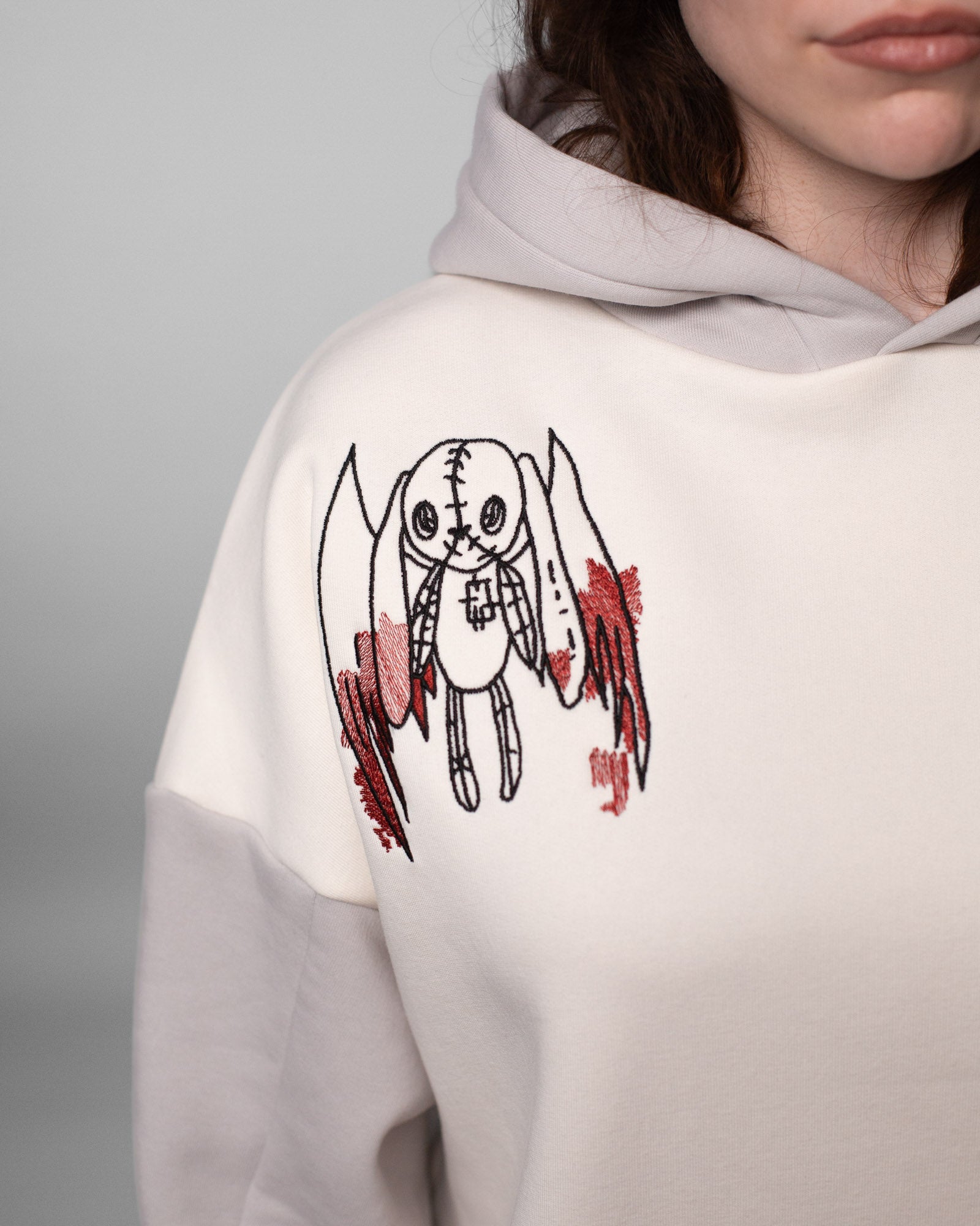 Nahaufnahme der gestickten Figur auf der linken Brust des Hoodies. Die Figur erinnert an ein geflügeltes Stofftier mit Nahtlinien, Knopfaugen und Kreuz auf der Brust. Um die Flügel herum sind blutrote Stickakzente gesetzt.