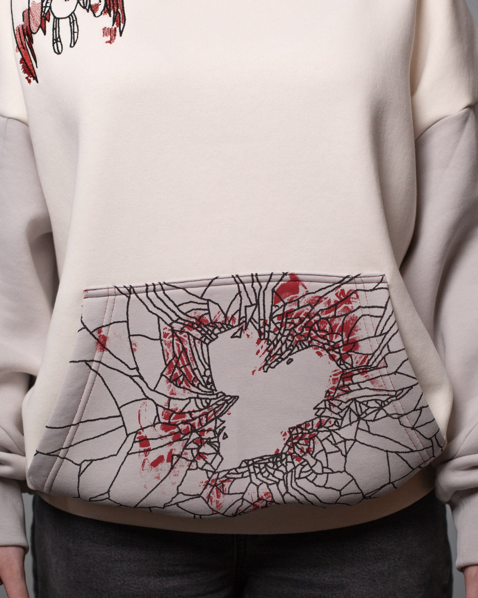 Detailansicht der Kängurutasche des Hoodies. Die Tasche ist mit einem schwarzen Spinnennetzmuster bedruckt, das eine zentrale Herzform aussparend umrahmt. Rote Farbeffekte suggerieren Blutspritzer.