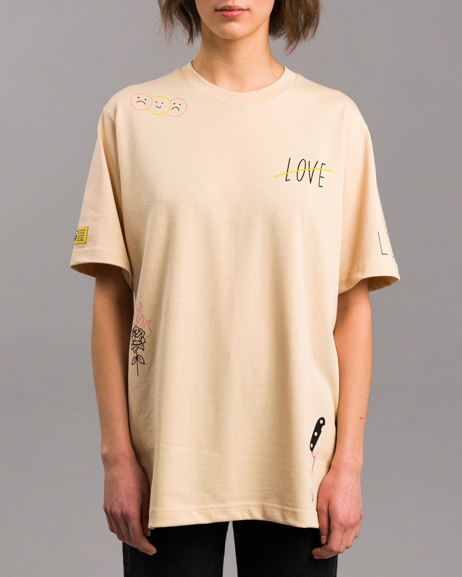 Frontalansicht des T-Shirts getragen von einer Frau. Links auf der Brust drei Smileys, rechts der durchgestrichene „LOVE“-Schriftzug. Am Saum eine brennende Rose und ein Messer mit gelb-pinken Blumen.
