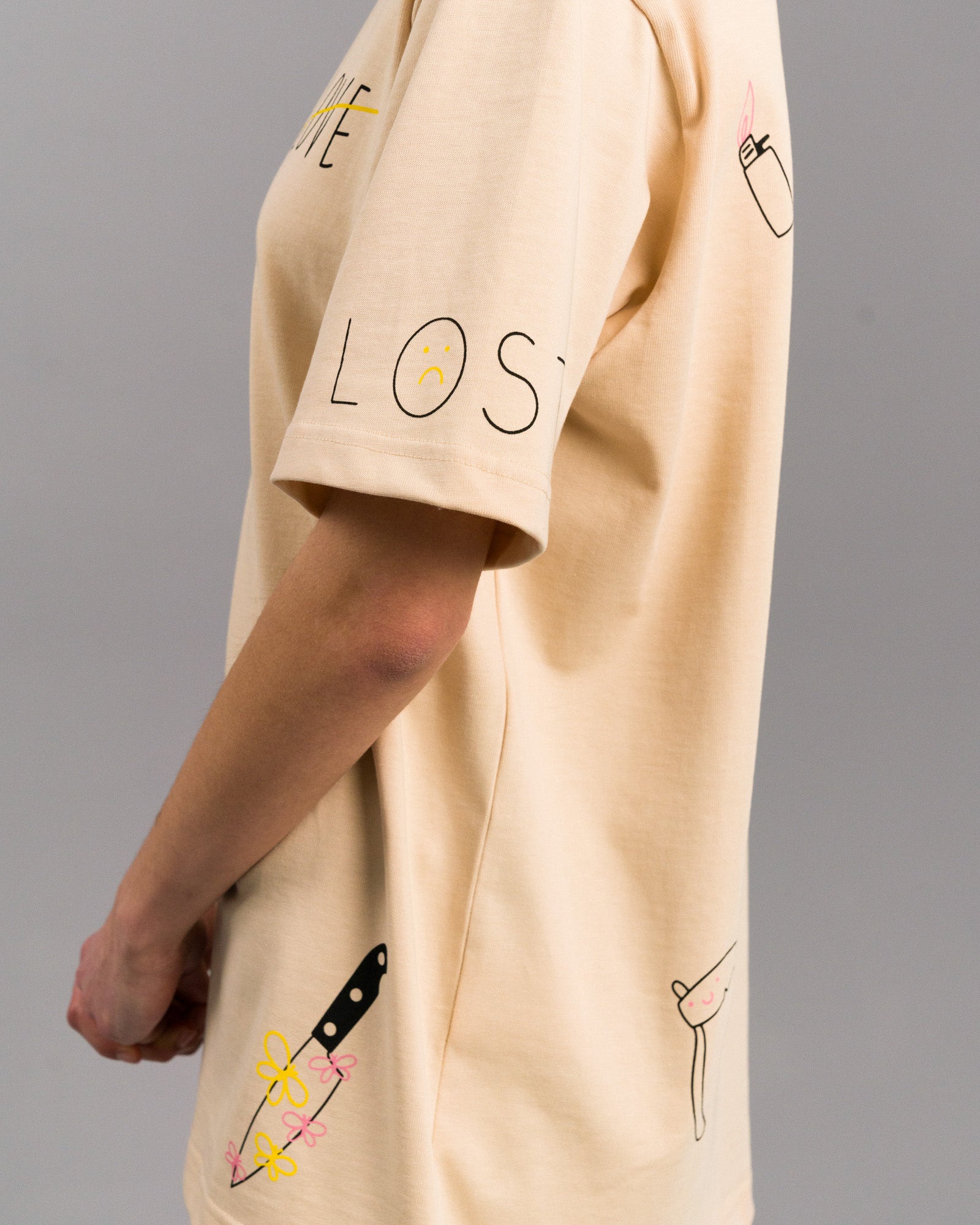 Seitenansicht des T-Shirts mit Schriftzug „LOST“ auf dem rechten Ärmel. Darunter ein Messer mit Blumen sowie eine kleine, gebogene Sense. Auf der Schulter ist ein rosa Feuerzeug aufgedruckt.