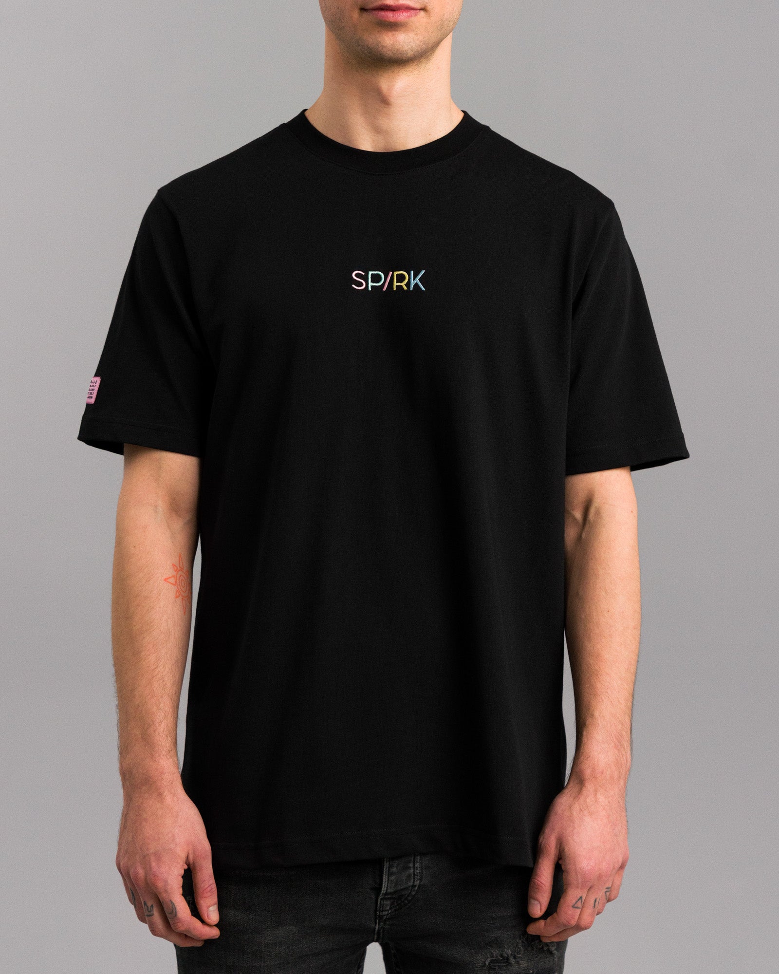 Schwarzes T-Shirt mit farbigem „SPARK“-Stick auf der Brust, getragen von einer Person mit dezenten Tattoos.