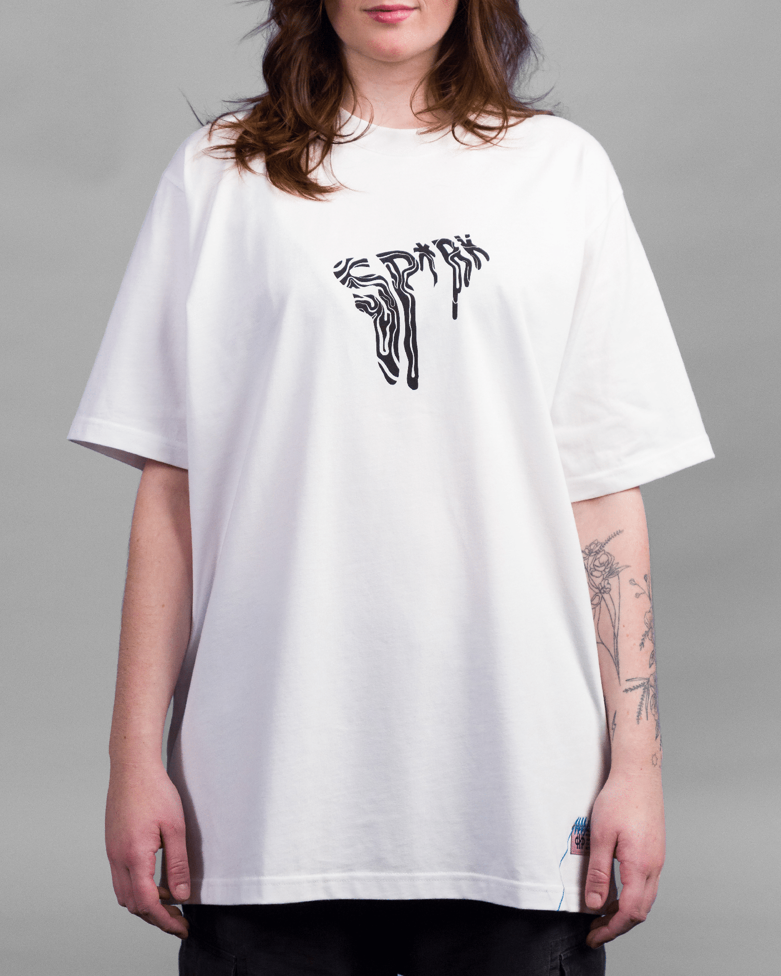 Weißes, oversized geschnittenes T-Shirt mit schwarzem Frontprint in fließender Liniengrafik, der das Wort „SP*RK“ stilisiert darstellt. Das Shirt endet locker über der Hüfte.