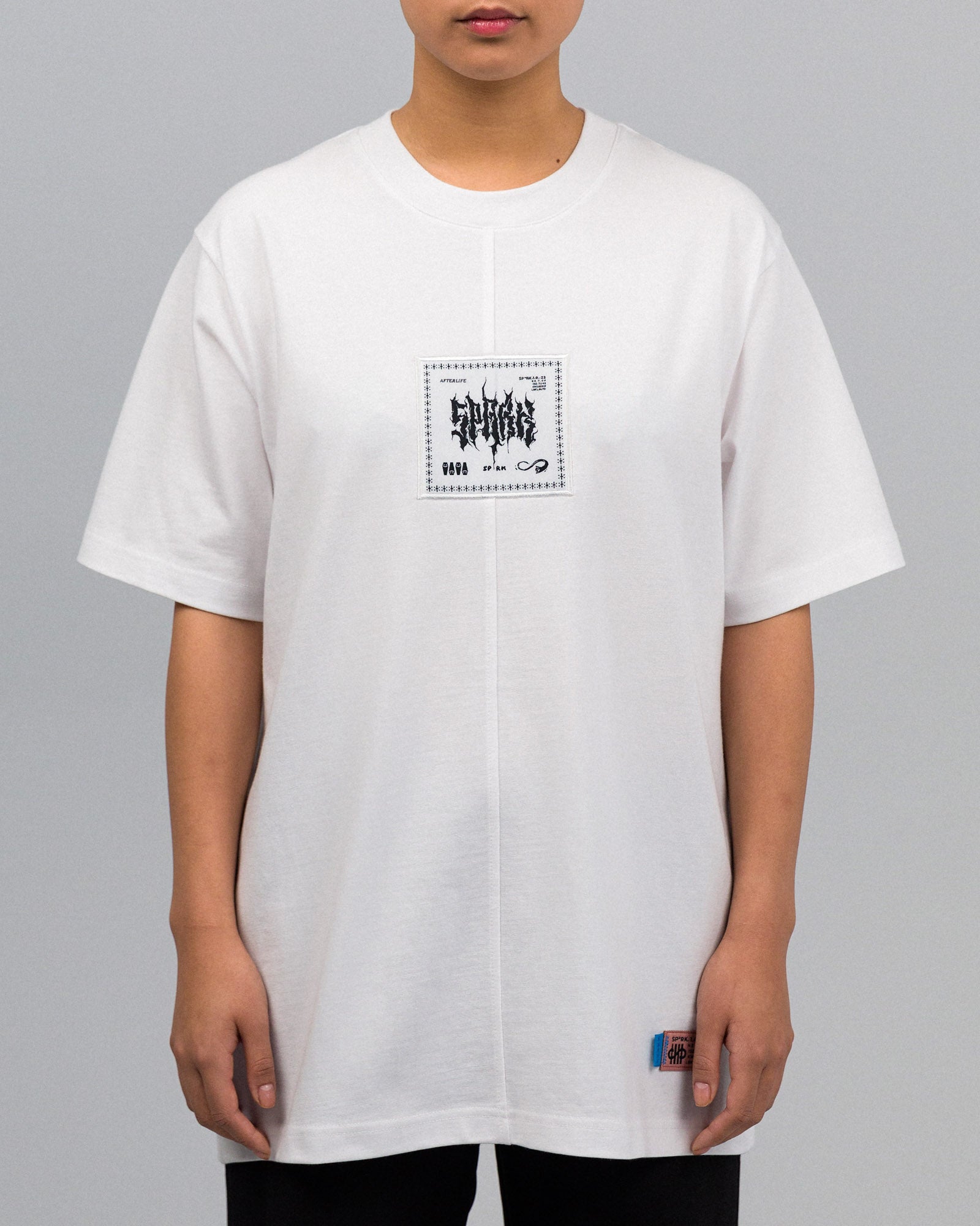 Frontalansicht einer Person im weißen T-Shirt. Das Shirt zeigt mittig auf der Brust ein quadratisches Patch mit der Aufschrift „AFTERLIFE“ und grafischen Elementen. Unten rechts ist das farbige Webetikett eingenäht.