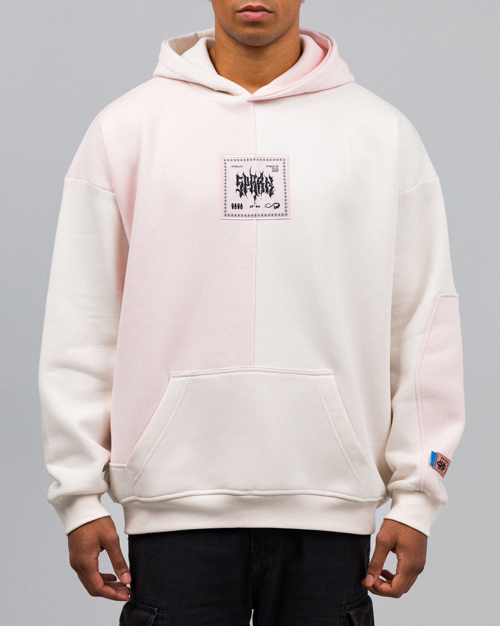 Locker geschnittener Hoodie mit zweifarbiger Vorderseite in Cremeweiß und Rosé. Auf der Brust ein quadratischer Aufnäher mit grafischem Schriftzug „SPARK“ und kleinen Symbolen. Kängurutasche im unteren Bereich.