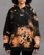 Schwarz-orangener Kapuzenpullover mit auffälligem Sprenkelmuster in Rasteroptik. Die orangefarbenen Punkte verlaufen diagonal über die Vorderseite. In Brusthöhe ist ein weißes, stilisiertes Logo mit gekreuzten Linien aufgestickt. Die Kapuze ist mit schwarzen Kordeln versehen, deren Enden leuchtend blau eingefärbt sind. Auf dem rechten Ärmelbündchen befindet sich ein gelbes Label mit blauer Aufschrift. Der Hoodie ist locker geschnitten und hat eine Kängurutasche auf Bauchhöhe.