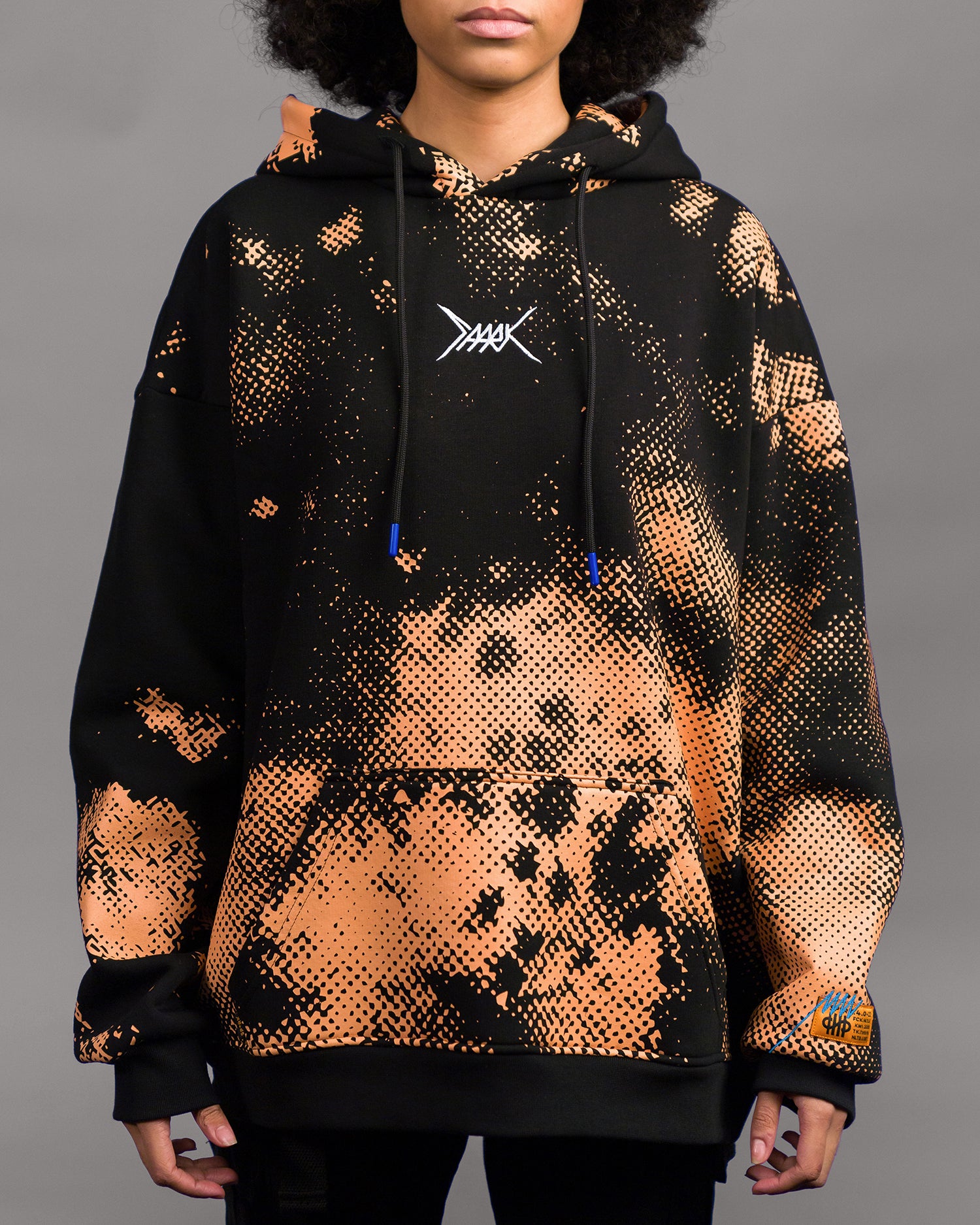 Schwarz-orangener Kapuzenpullover mit auffälligem Sprenkelmuster in Rasteroptik. Die orangefarbenen Punkte verlaufen diagonal über die Vorderseite. In Brusthöhe ist ein weißes, stilisiertes Logo mit gekreuzten Linien aufgestickt. Die Kapuze ist mit schwarzen Kordeln versehen, deren Enden leuchtend blau eingefärbt sind. Auf dem rechten Ärmelbündchen befindet sich ein gelbes Label mit blauer Aufschrift. Der Hoodie ist locker geschnitten und hat eine Kängurutasche auf Bauchhöhe.