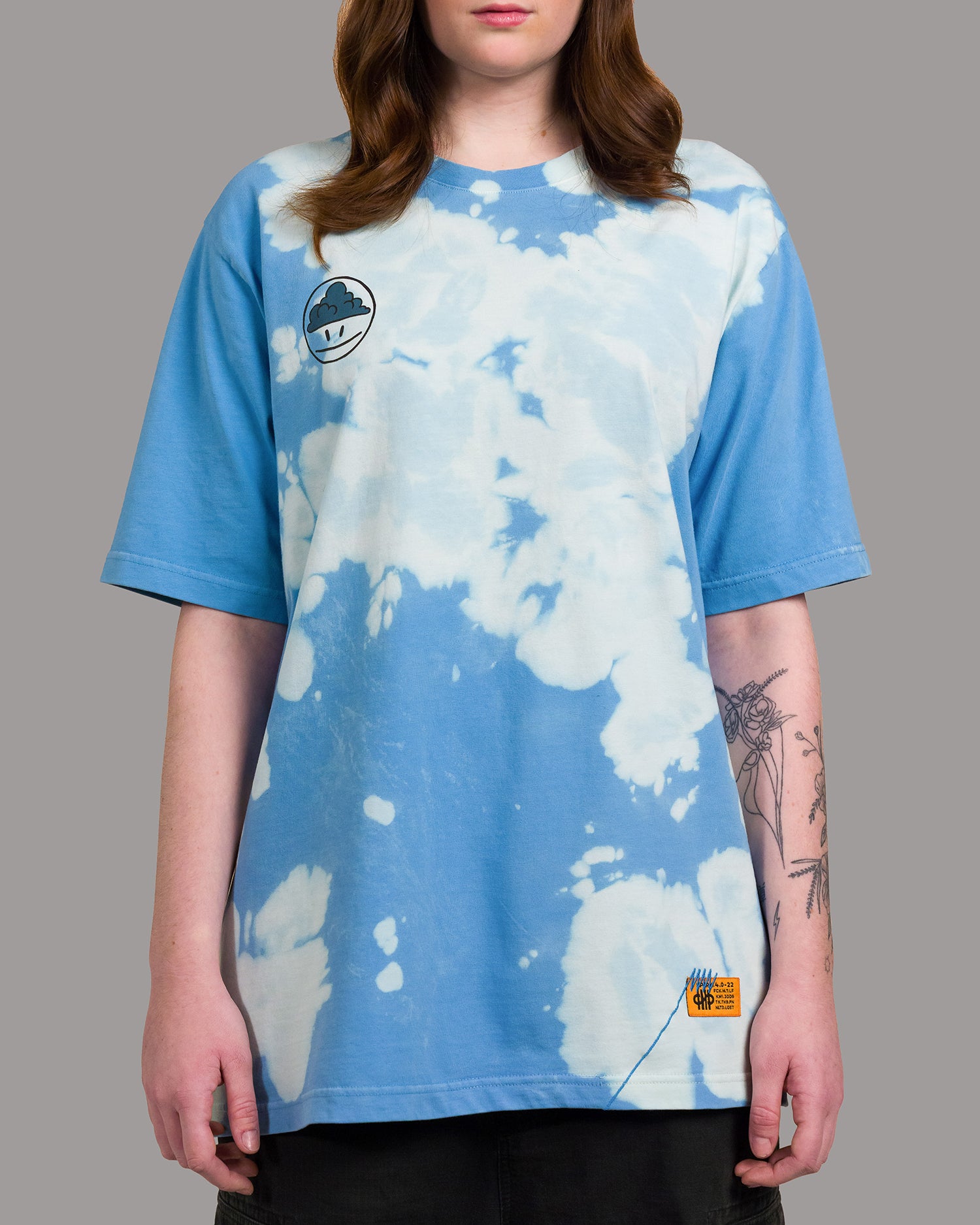 T-Shirt mit wolkenähnlichem Batikmuster in Hellblau und Weiß. Auf der linken Brust ist ein aufgedrucktes Smiley-Motiv mit dunkler Wolke im Kopfbereich zu sehen. Am unteren Saum befindet sich ein orangefarbenes Label mit blauen Ziernähten.
