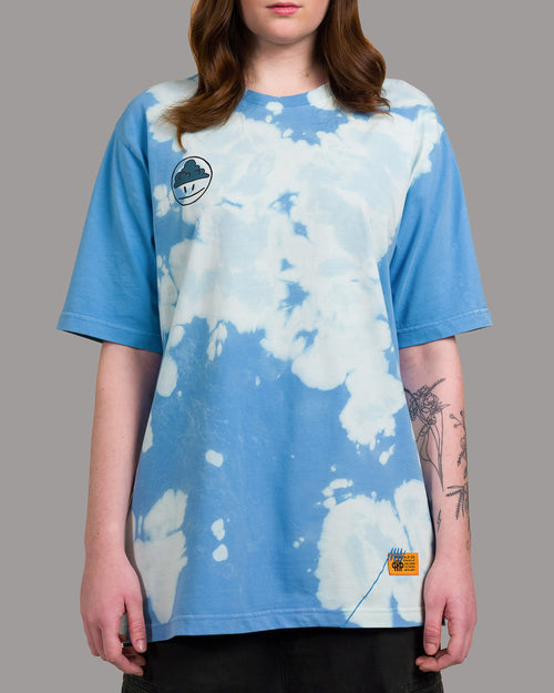 T-Shirt mit wolkenähnlichem Batikmuster in Hellblau und Weiß. Auf der linken Brust ist ein aufgedrucktes Smiley-Motiv mit dunkler Wolke im Kopfbereich zu sehen. Am unteren Saum befindet sich ein orangefarbenes Label mit blauen Ziernähten.