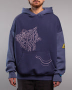 Frontalansicht eines dunkelblauen Hoodies mit aufgedrucktem Linienmotiv auf der Brust, das eine abstrakte, organisch geschwungene Form zeigt. Unten auf der Kängurutasche verlaufen drei wellenartige Linien nach rechts. Die Ärmel sind in leicht hellerem Blau abgesetzt, am linken Oberarm befindet sich ein gelb-schwarzes, dreieckiges Labelpatch.