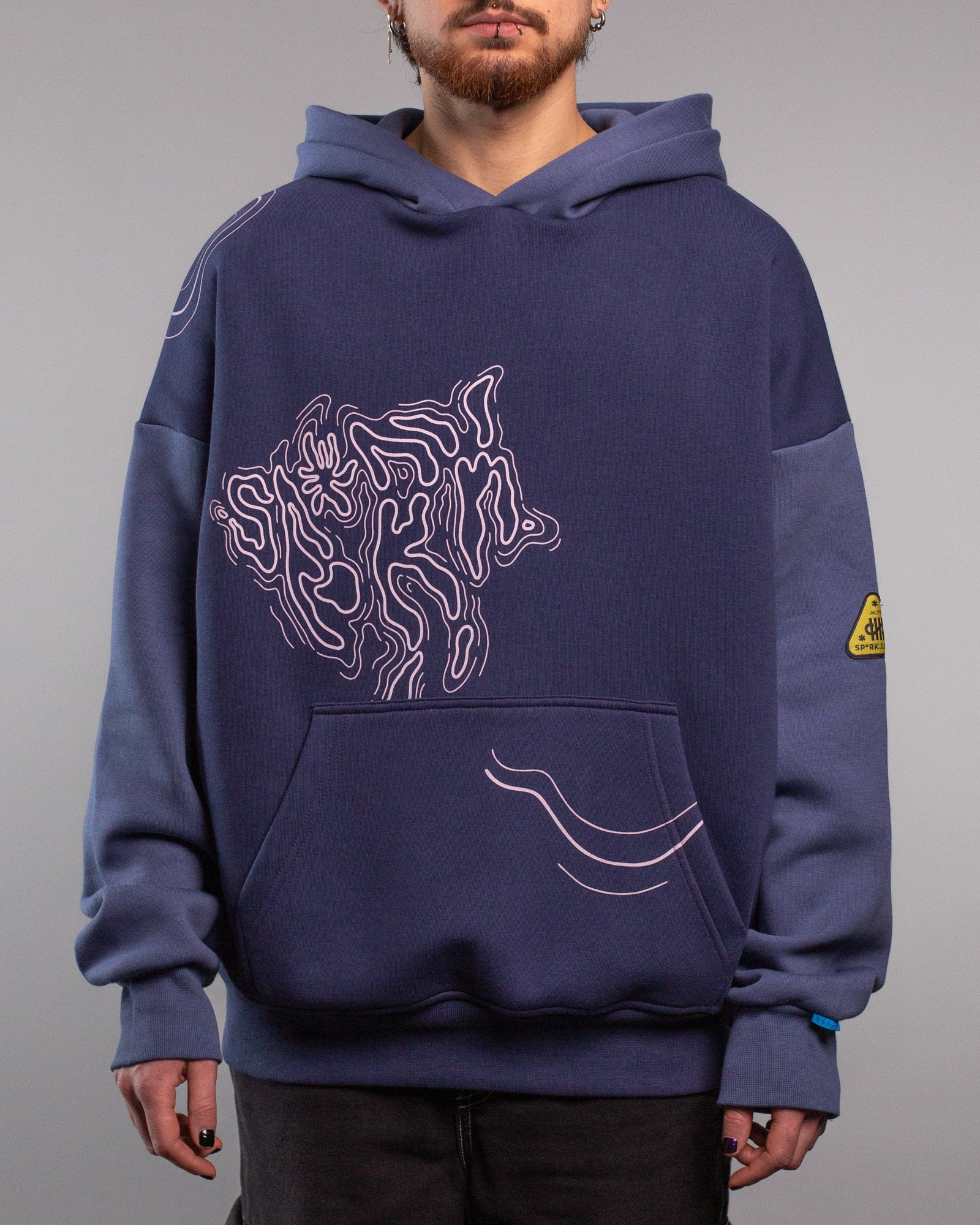 Frontalansicht eines dunkelblauen Hoodies mit aufgedrucktem Linienmotiv auf der Brust, das eine abstrakte, organisch geschwungene Form zeigt. Unten auf der Kängurutasche verlaufen drei wellenartige Linien nach rechts. Die Ärmel sind in leicht hellerem Blau abgesetzt, am linken Oberarm befindet sich ein gelb-schwarzes, dreieckiges Labelpatch.
