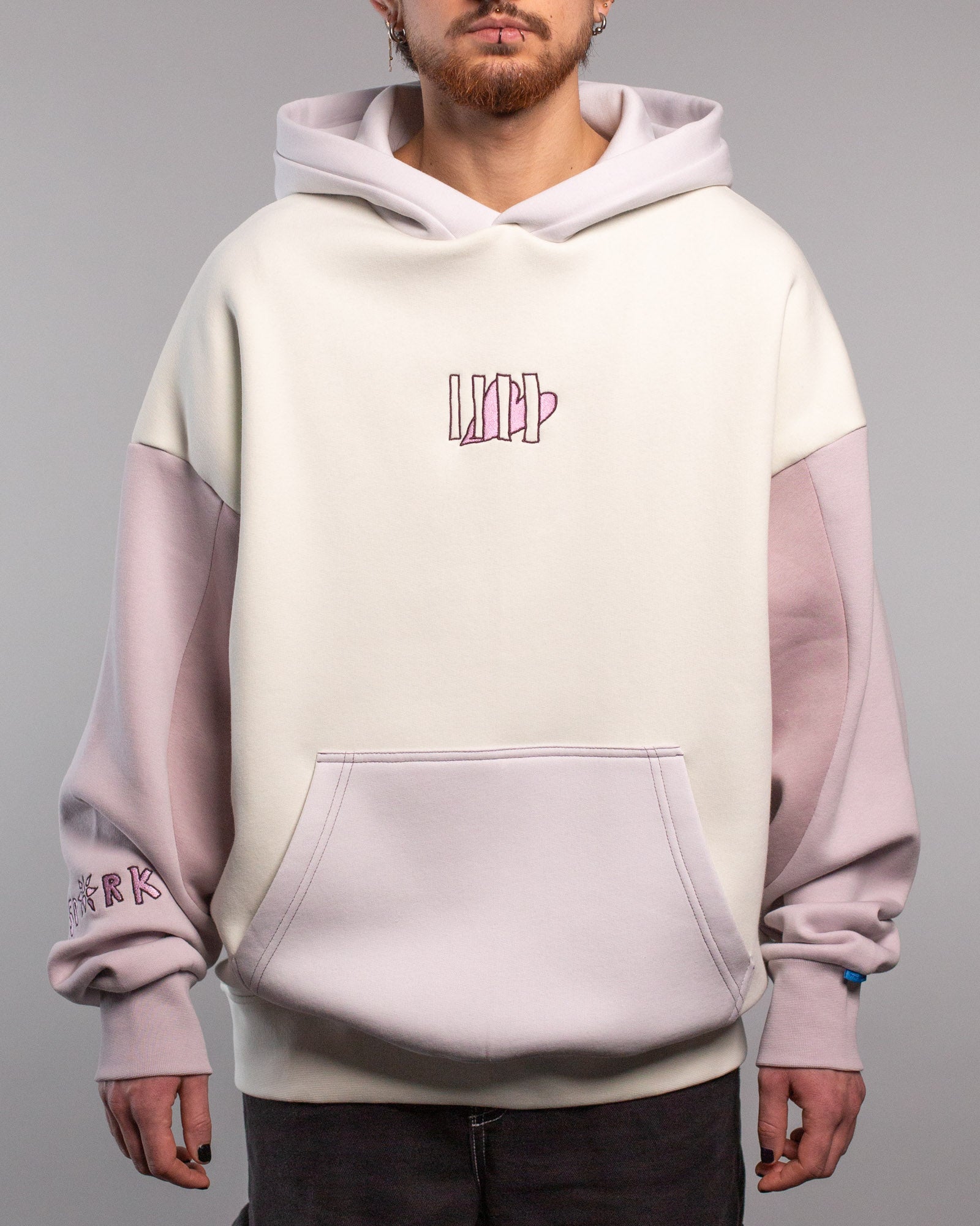 Cremefarbener Hoodie mit fliederfarbenen Ärmeln, getragen von vorn gezeigt. Auf der Brust ist ein rosa Herzmotiv zu sehen, das von fünf senkrechten Linien in dunklem Lila umgeben ist. Die Ärmel sind im Farbblockstil gehalten, die Kapuze ist ebenfalls fliederfarben.