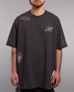 Frontalansicht eines schwarzen T-Shirts mit bunten grafischen Prints. Auf der linken Schulter drei Smiley-Gesichter (traurig, fröhlich, traurig), auf der rechten Brust das durchgestrichene Wort „LOVE“ in Weiß mit gelbem Strich. Unten links eine weiße Rose mit pinken Flammen, unten rechts ein Messer mit floraler Verzierung.