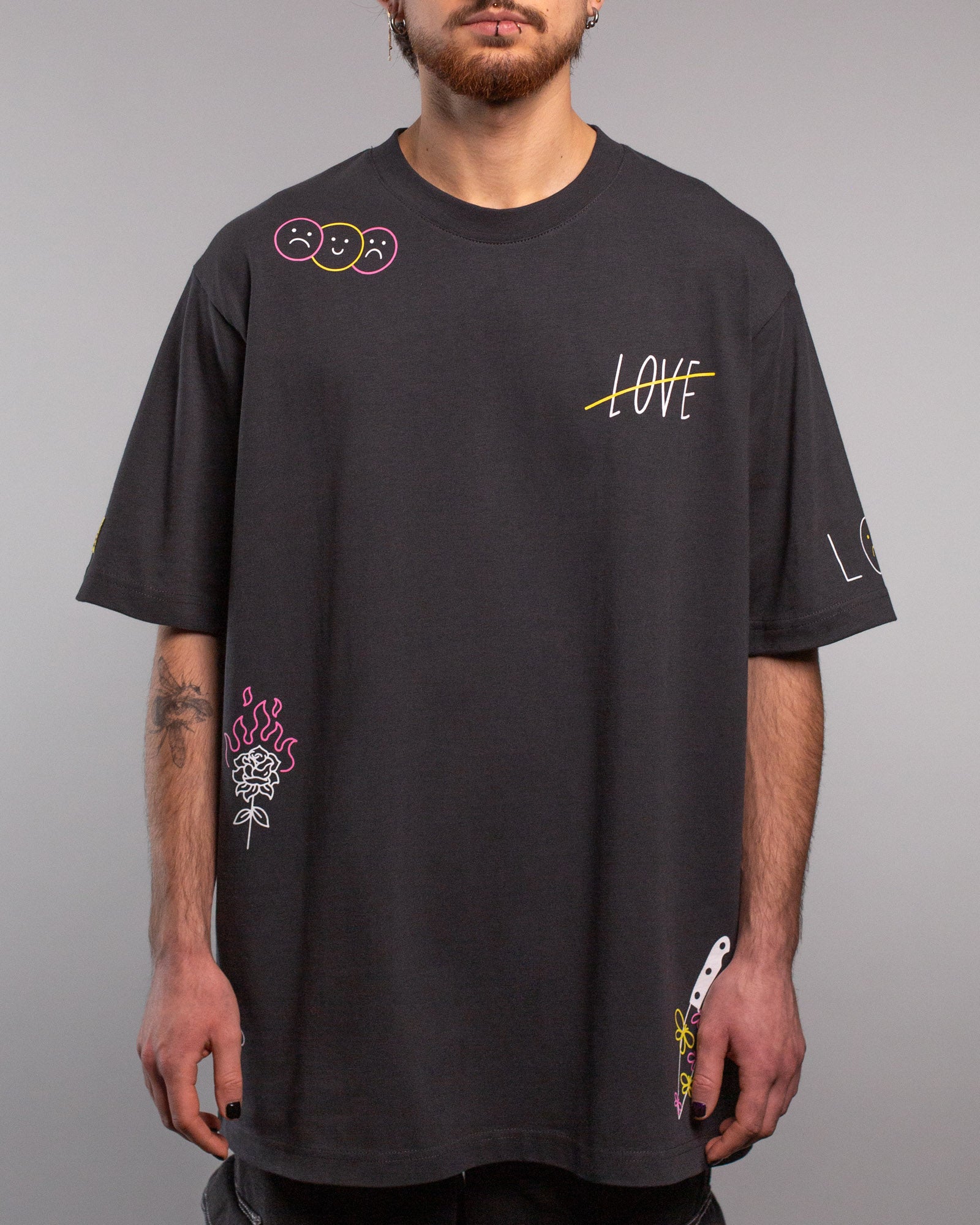 Frontalansicht eines schwarzen T-Shirts mit bunten grafischen Prints. Auf der linken Schulter drei Smiley-Gesichter (traurig, fröhlich, traurig), auf der rechten Brust das durchgestrichene Wort „LOVE“ in Weiß mit gelbem Strich. Unten links eine weiße Rose mit pinken Flammen, unten rechts ein Messer mit floraler Verzierung.