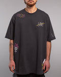 Frontalansicht eines schwarzen T-Shirts mit bunten grafischen Prints. Auf der linken Schulter drei Smiley-Gesichter (traurig, fröhlich, traurig), auf der rechten Brust das durchgestrichene Wort „LOVE“ in Weiß mit gelbem Strich. Unten links eine weiße Rose mit pinken Flammen, unten rechts ein Messer mit floraler Verzierung.