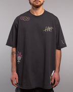 Frontalansicht eines schwarzen T-Shirts mit bunten grafischen Prints. Auf der linken Schulter drei Smiley-Gesichter (traurig, fröhlich, traurig), auf der rechten Brust das durchgestrichene Wort „LOVE“ in Weiß mit gelbem Strich. Unten links eine weiße Rose mit pinken Flammen, unten rechts ein Messer mit floraler Verzierung.