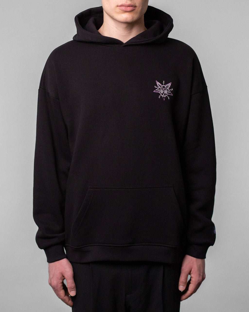 Schwarzer Hoodie in Frontalansicht, getragen von einer Person. Auf der linken Brustseite befindet sich ein kleines, lilafarbenes Grafikmotiv in Sternform mit einem stilisierten Ziegenkopf, spiralförmigen Augen und dem Schriftzug „SPARK“ in den Sternzacken.