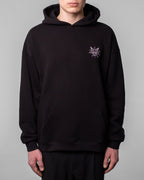 Schwarzer Hoodie in Frontalansicht, getragen von einer Person. Auf der linken Brustseite befindet sich ein kleines, lilafarbenes Grafikmotiv in Sternform mit einem stilisierten Ziegenkopf, spiralförmigen Augen und dem Schriftzug „SPARK“ in den Sternzacken.