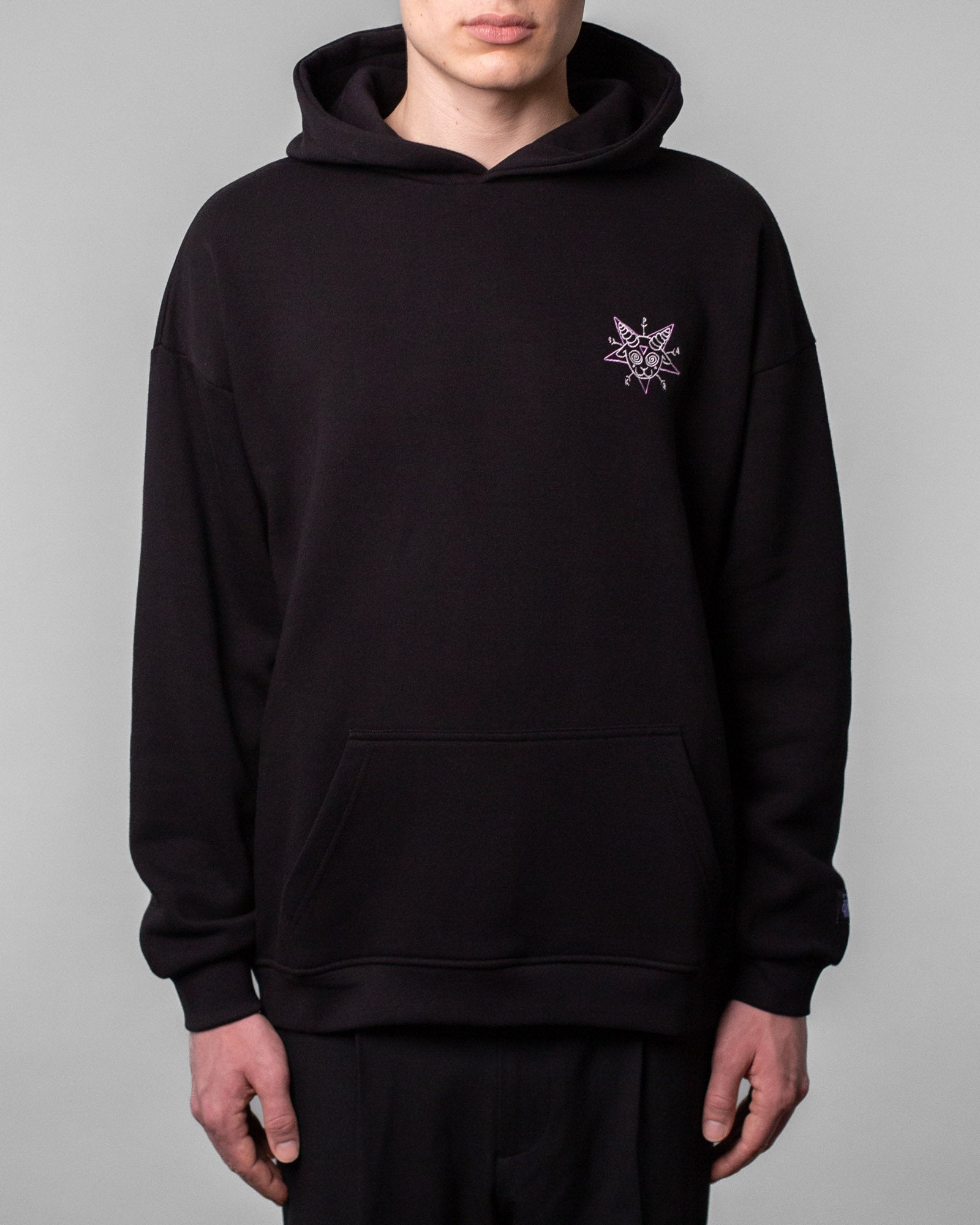 Schwarzer Hoodie in Frontalansicht, getragen von einer Person. Auf der linken Brustseite befindet sich ein kleines, lilafarbenes Grafikmotiv in Sternform mit einem stilisierten Ziegenkopf, spiralförmigen Augen und dem Schriftzug „SPARK“ in den Sternzacken.