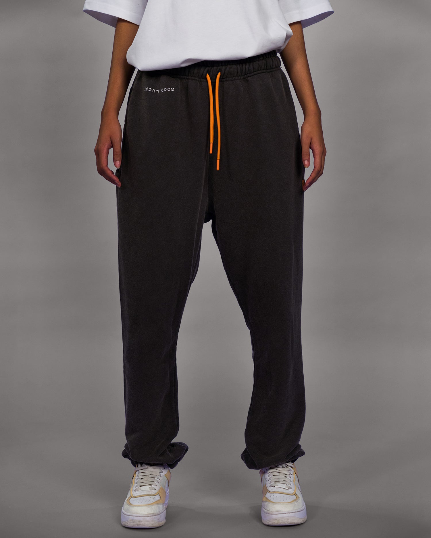 Schwarze Jogginghose mit orangefarbenem Kordelzug und weißem, umgedrehtem „good luck“-Schriftzug unter dem Bund auf der linken Seite. Die Hose hat einen weiten, lockeren Schnitt und wird mit weißen Sneakern getragen. Hintergrund grau.

