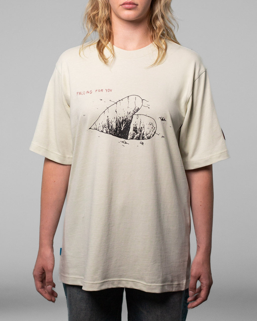 Eine Person trägt ein beigefarbenes T-Shirt mit schwarzem Grafikprint auf der Vorderseite. Das Motiv zeigt ein großes, halbiertes Herz mit strukturierter Füllung und feinen Linien, die an Pflanzen oder Risse erinnern. Darüber steht der rote Schriftzug „FALLING FOR YOU“. Das Shirt hat eine lockere Passform mit kurzen Ärmeln und Rundhalsausschnitt. Der Fokus liegt auf dem illustrativen Frontdesign und dem Schriftzug.