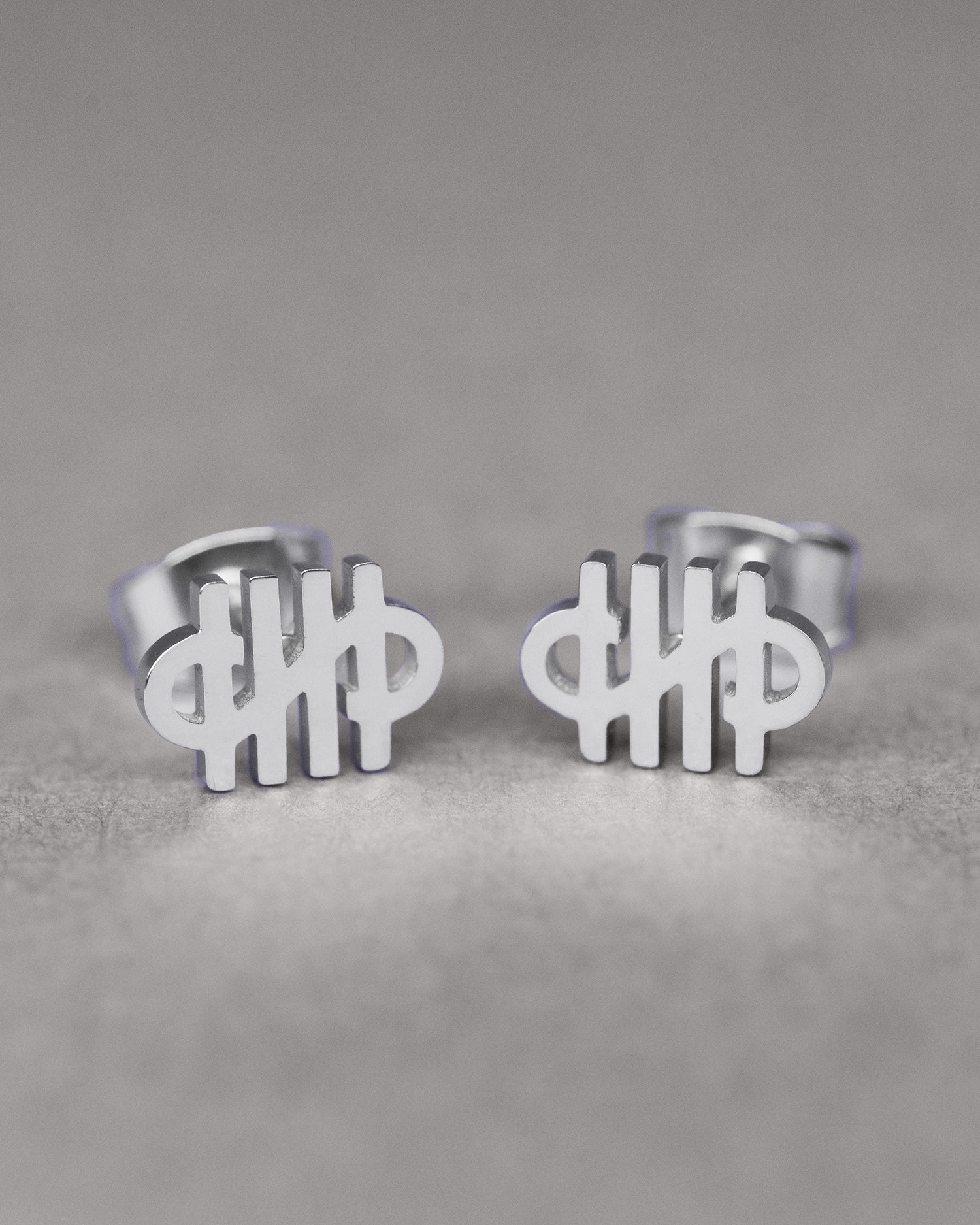 Nahaufnahme eines Paars silberner Ohrstecker in Form eines grafischen Logos mit vier vertikalen Linien und einer geschwungenen Form, die an ein Monogramm erinnert. Präsentiert auf grauer Oberfläche.