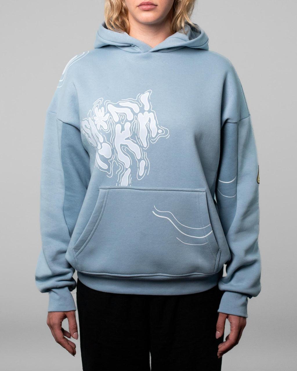 Eine Person trägt einen hellblauen Kapuzenpullover mit weißem, abstraktem Linienmotiv auf der linken Brustseite und entlang der Kängurutasche. Der Hoodie hat eine weite Passform mit breiten Bündchen an Ärmeln und Saum. Der Fokus liegt auf dem grafischen Frontdesign und der hochwertigen Verarbeitung des Stoffes. Der Hintergrund ist neutral grau gehalten.