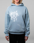 Eine Person trägt einen hellblauen Kapuzenpullover mit weißem, abstraktem Linienmotiv auf der linken Brustseite und entlang der Kängurutasche. Der Hoodie hat eine weite Passform mit breiten Bündchen an Ärmeln und Saum. Der Fokus liegt auf dem grafischen Frontdesign und der hochwertigen Verarbeitung des Stoffes. Der Hintergrund ist neutral grau gehalten.
