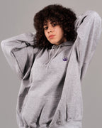 Grauer Kapuzenpullover in weiter Passform mit violettem Flammen-Logo auf der linken Brust. Der Hoodie besitzt eine Kängurutasche und helle Kordeln an der Kapuze. Das Produkt wird von einer Person mit lockigem Haar getragen, die die Hände hinter dem Kopf verschränkt hat.
