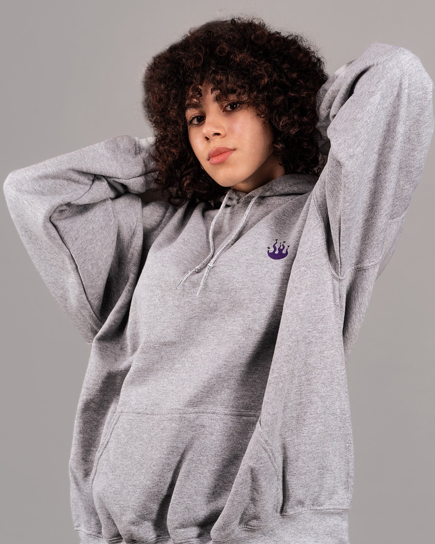 Grauer Kapuzenpullover in weiter Passform mit violettem Flammen-Logo auf der linken Brust. Der Hoodie besitzt eine Kängurutasche und helle Kordeln an der Kapuze. Das Produkt wird von einer Person mit lockigem Haar getragen, die die Hände hinter dem Kopf verschränkt hat.
