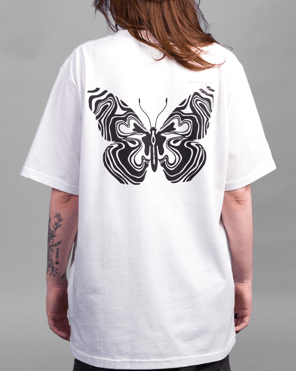 Auf dem Rücken des weißen T-Shirts befindet sich ein großes, symmetrisches Schmetterlingsmotiv aus schwarzen, psychedelisch anmutenden Linien. Rechts oben ist ein kleiner Textprint in Rosa und Schwarz sichtbar.