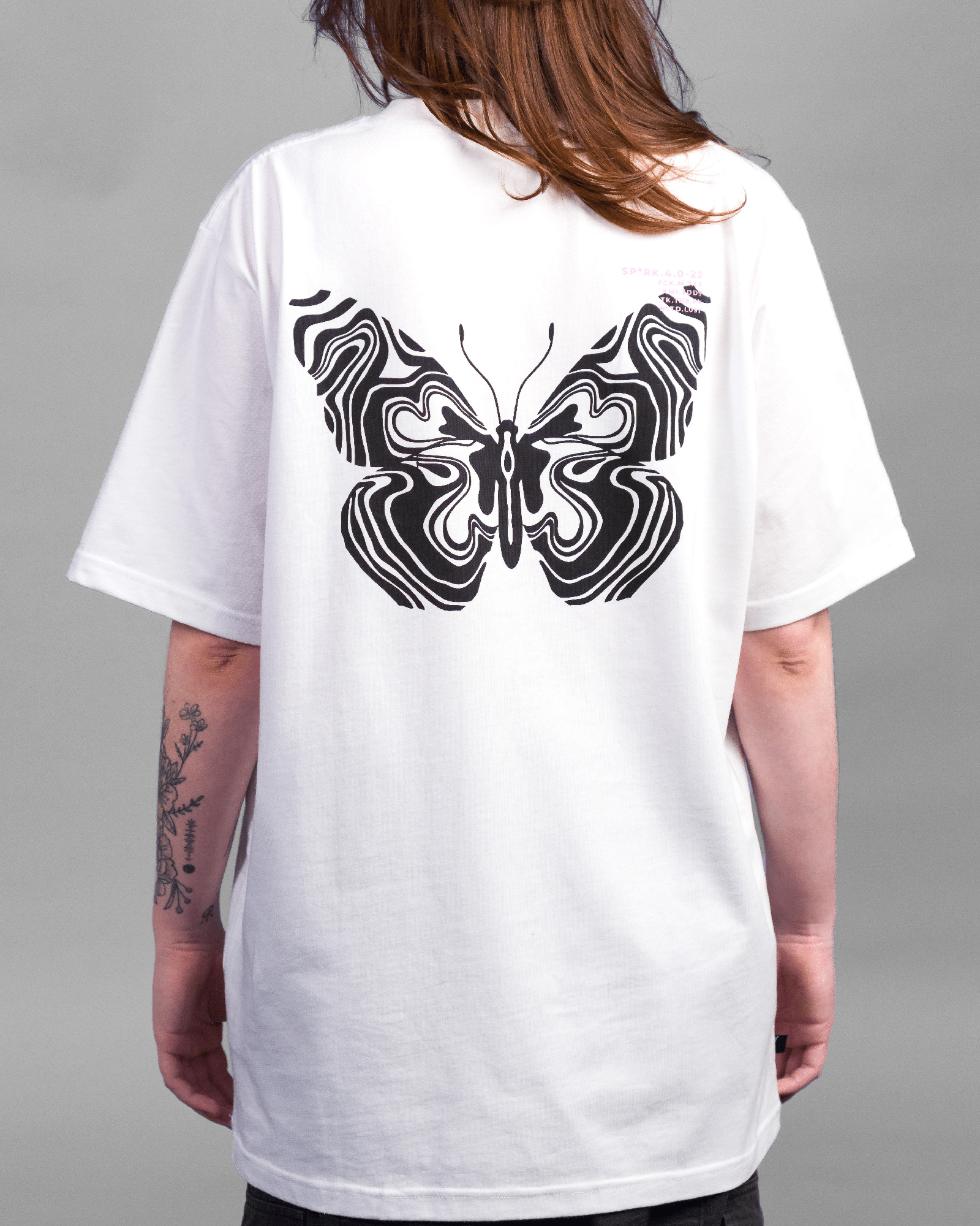 Auf dem Rücken des weißen T-Shirts befindet sich ein großes, symmetrisches Schmetterlingsmotiv aus schwarzen, psychedelisch anmutenden Linien. Rechts oben ist ein kleiner Textprint in Rosa und Schwarz sichtbar.