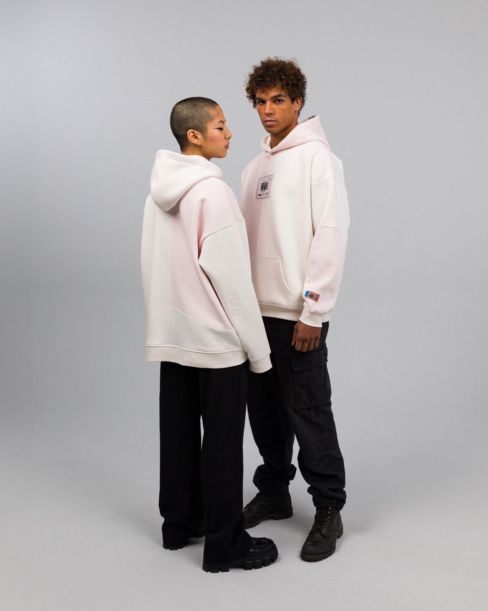 Zwei Personen tragen einen zweifarbigen Hoodie in Cremeweiß und hellem Rosa. Die Vorderseite zeigt einen zentralen Patch mit grafischem Print. Der Hoodie hat eine Kängurutasche und ist locker geschnitten.