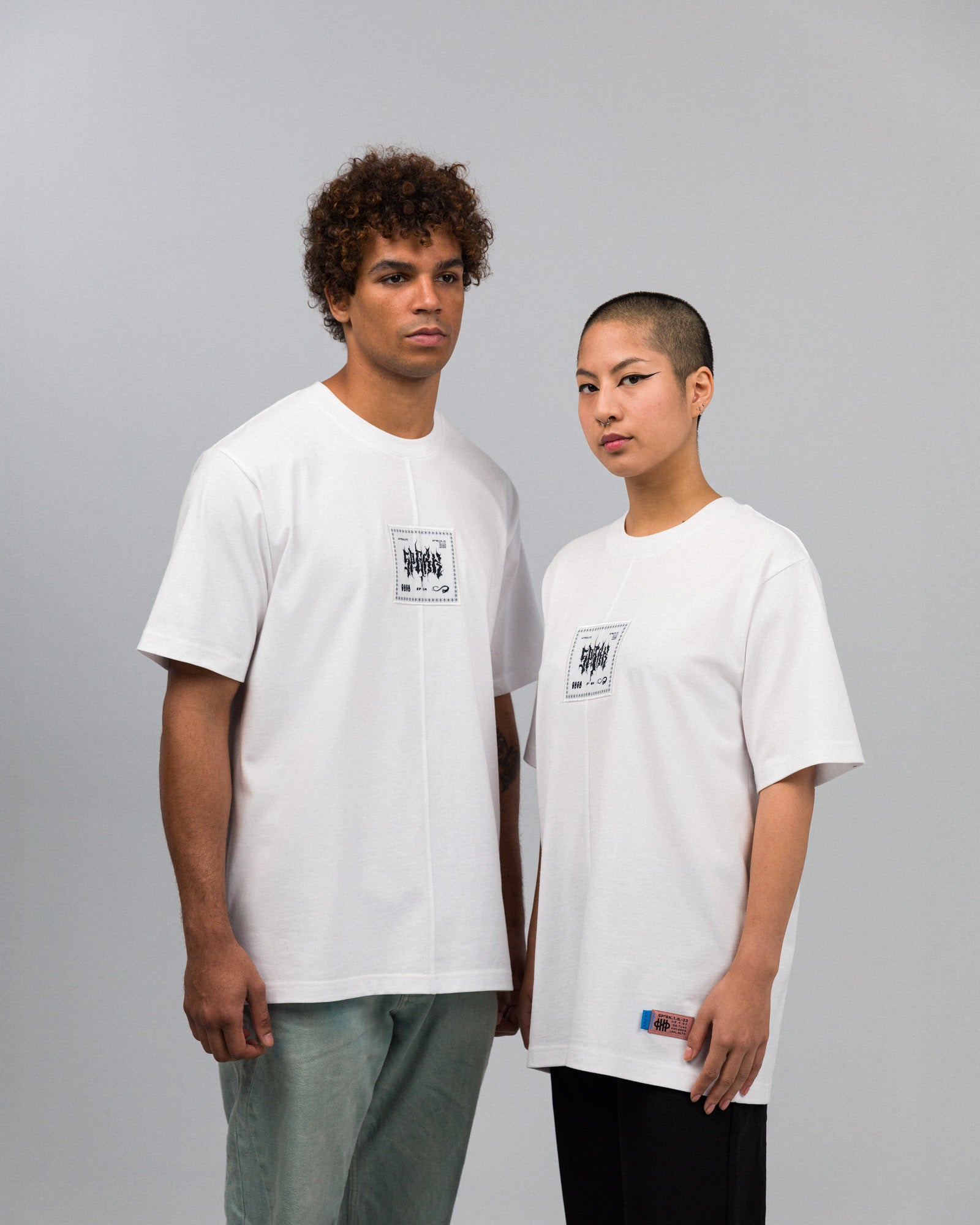 Zwei Personen tragen weiße T-Shirts mit mittigem, quadratischem Frontprint in Schwarz mit grafischer Typografie und Symbolen. Das Shirt ist locker geschnitten, mit sichtbarer vertikaler Mittelnaht. Die linke Person kombiniert es mit mintgrüner Hose, die rechte mit schwarzer Hose.
