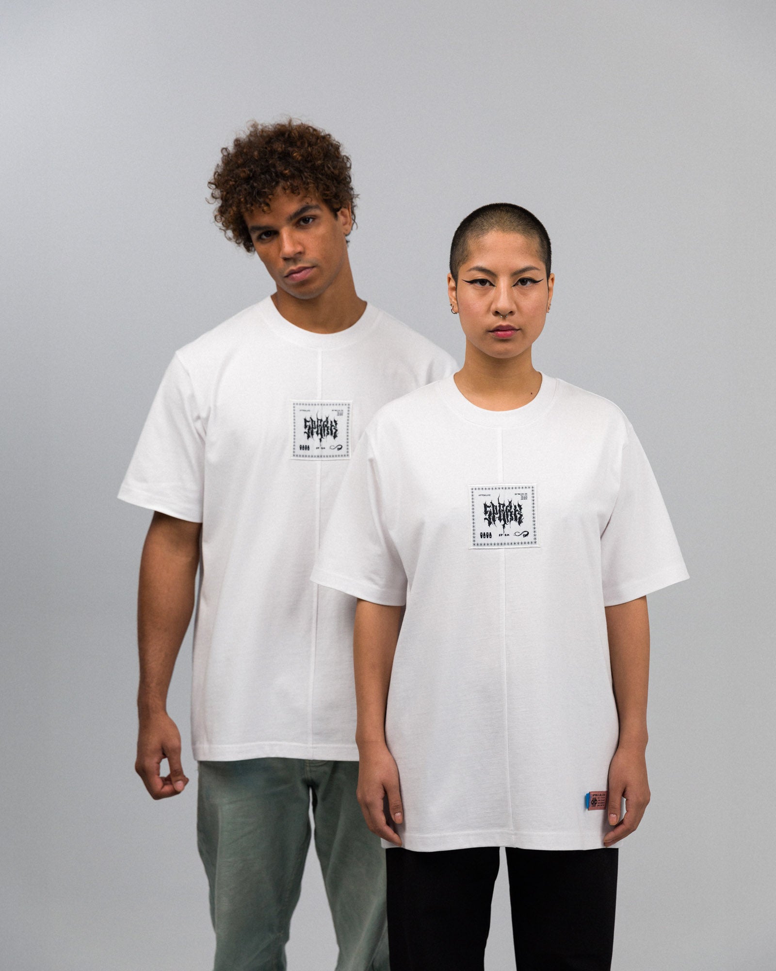 Die beiden Models stehen frontal zur Kamera und tragen identische weiße T-Shirts mit schwarzem, quadratischem Frontprint. Die Naht verläuft mittig von Kragen bis Saum. Am Saum des rechten Shirts ist ein auffälliges gewebtes Etikett sichtbar.