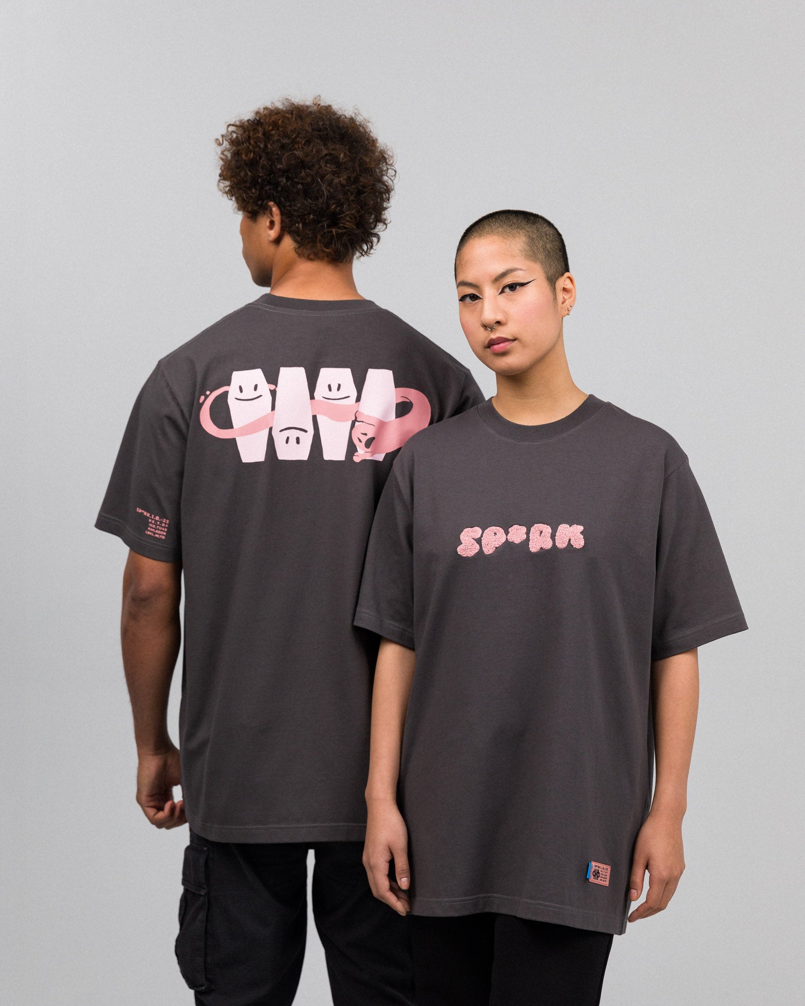 Zwei Personen tragen ein dunkelgraues T-Shirt mit rosa Aufdrucken. Die Person vorn zeigt den Frontprint „SP*RK“ in plüschiger, rosa Typografie auf Brusthöhe. Die rückwärtige Person zeigt den großflächigen Rückenprint mit vier stilisierten Särgen mit Gesichtern und einem rosa Band, das sich schlaufenförmig durch die Motive windet.