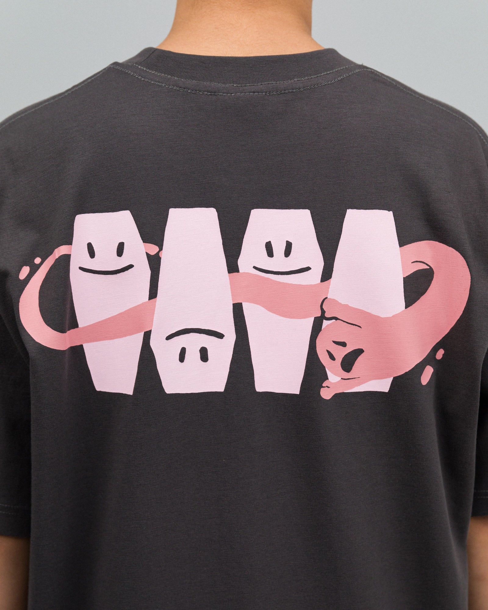 Detailaufnahme des Rückenprints auf dem dunkelgrauen T-Shirt: Vier rosa Särge mit unterschiedlichen Gesichtsausdrücken stehen nebeneinander, durchzogen von einem rosa, schlangenartigen Band mit Katzengesicht und ausgestreckter Pfote.