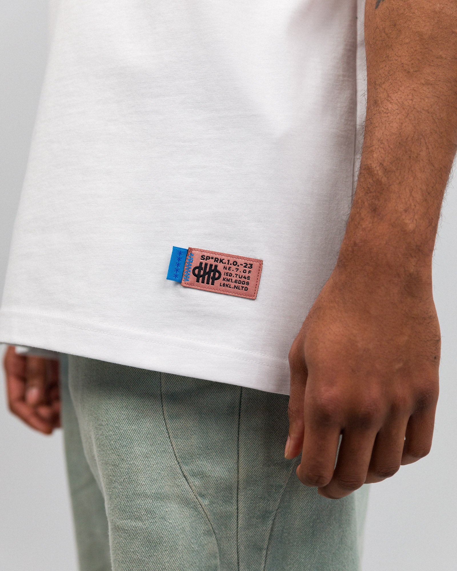 Nahaufnahme des gewebten Labels am Saum des weißen T-Shirts. Das Label ist rechteckig, rot-blau mit schwarzem Text. Es enthält alphanumerische Codes und das Logo der Marke „SP*RK“.