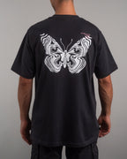 Rückenansicht desselben schwarzen T-Shirts mit großflächigem, weißem Schmetterlingsprint in marmorierter Liniengrafik. Rechts oben ist ein kleiner rosa Textprint mit Zahlen- und Buchstabenkombinationen sichtbar.
